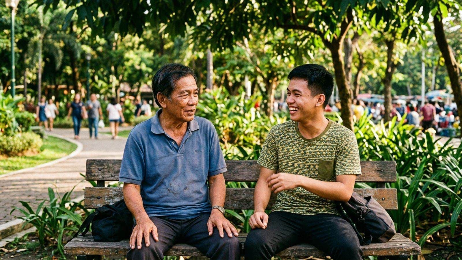 An older man and a young man laugh together on a wooden park bench under shade trees