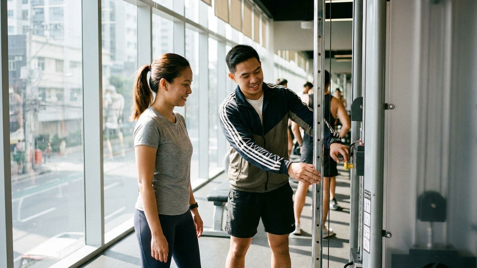 A coach shows a smiling beginner how to set the weight on a cable machine, beside the tall windows of a city gym